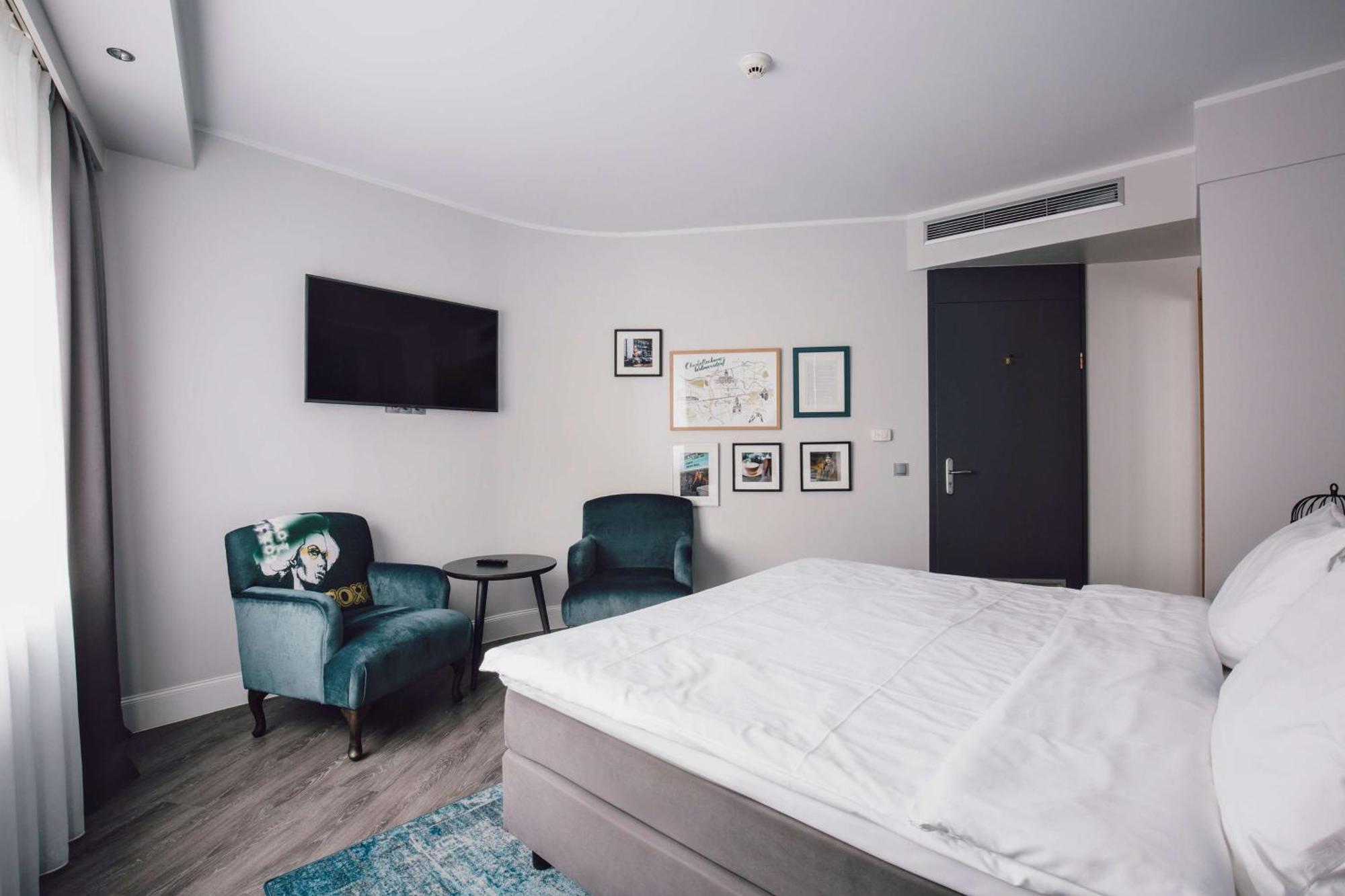 Hotel Berlin, Berlin, A Member Of Radisson Individuals Buitenkant foto The photo depicts a modern hotel room with a minimalist design. There is a large bed with white linens and a grey headboard. To the right, two plush, teal armchairs are positioned next to a small round table. A wall-mounted television is visible abov