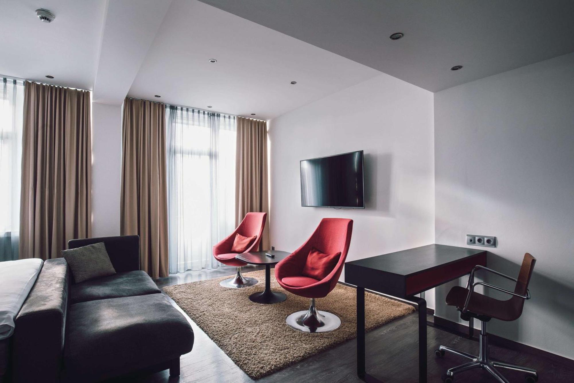 Hotel Berlin, Berlin, A Member Of Radisson Individuals Buitenkant foto The photo shows a modern living space with a stylish design. It features a dark gray sectional sofa, two red accent chairs around a circular black table, and a wall-mounted flat-screen television. There is a plush area rug on the floor, and large win